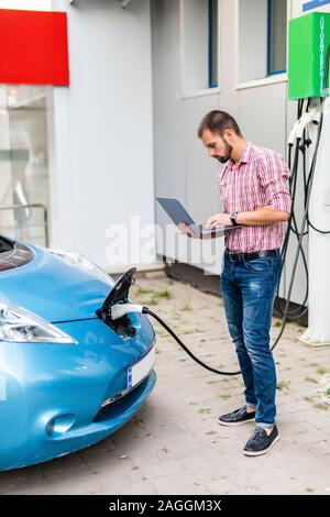 Beau jeune homme avec un ordinateur portable près de voiture électrique de charge Banque D'Images