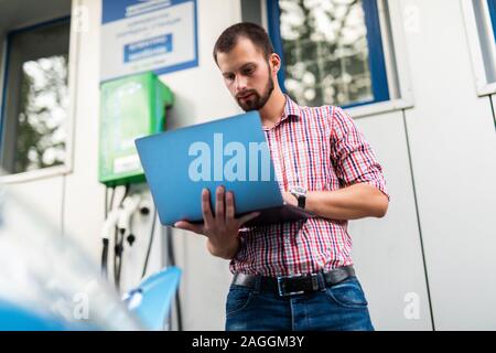 Beau jeune homme avec un ordinateur portable près de voiture électrique de charge Banque D'Images