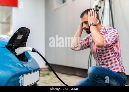 Bel homme de brancher le câble à la voiture électrique avec les mains sur la tête choquée. Banque D'Images