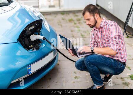 Bel homme avec un ordinateur portable près de voiture électrique de charge Banque D'Images