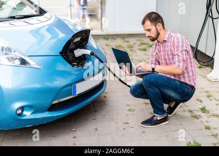 Bel homme avec un ordinateur portable près de voiture électrique de charge Banque D'Images