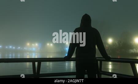 Une silhouette encapuchonnée silhouetté contre l'éclairage de rue à côté d'une rivière. Sur une mystérieuse hivers brumeux, nuit Banque D'Images