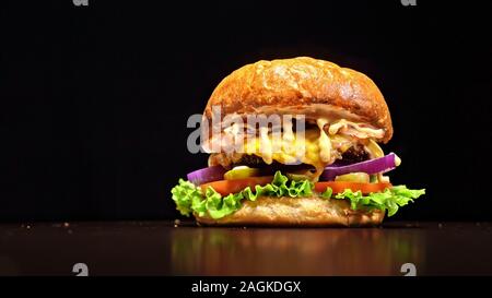 Burger d'artisanat est cuisson sur fond noir. Composé : sauce, salade, tomate, oignon rouge, cornichon, fromage, bacon, pain et viande marbrée de boeuf. Banque D'Images