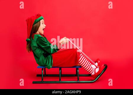 Toute la longueur du côté de la photo de profil elf excité kid en chapeau à tête rouge d'une longueur de coupe de chiens hurlant costume wearing cap sur rouge isolé Banque D'Images