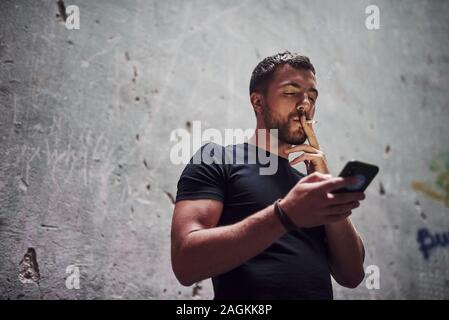 Par messagerie à l'aide de téléphone. Portrait d'un homme en chemise noire fumeurs sur l'arrière-plan de vieux mur fissuré Banque D'Images