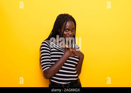 Portrait de heureux touché young African American Woman sourit doucement, se sent heureux, porte haut rayé et casquettes, isolé sur fond jaune Banque D'Images