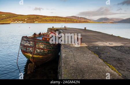 La dégradation de l'épave d'un petit bateau amarré a menti à côté d'une pierre jetée de Port Bannatyne sur l'île de Bute, avec les montagnes des hautes terres de Scotl Banque D'Images