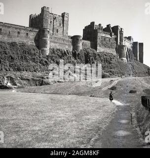 Années 1950, historique, une dame marchant le long d'un chemin dans le domaine sous le château de Bamburgh construit sur une colline, Northumberland, Angleterre, Royaume-Uni. Banque D'Images