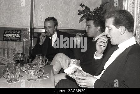 Années 1970, historique, trois hommes assis dans un coin d'un bar ou d'un pub mangeant des chips et profitant d'un tankard de bière, Angleterre, Royaume-Uni. Un signe pour la dernière boisson, une «boule de neige» peut être vu sur le manteau de la cheminée. Un cocktail, une boule de neige a été faite avec advocaat et limonade. Banque D'Images