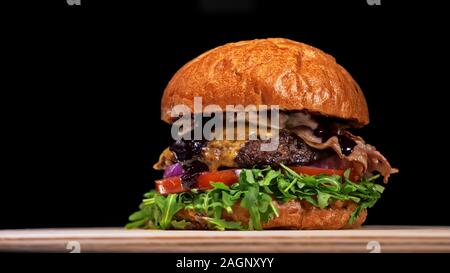 Burger d'artisanat est cuisson sur fond noir. Composé : sauce, roquette, tomate, oignon, bacon, fromage ricotta, sauce au cassis, pain d'air et en viande. Banque D'Images