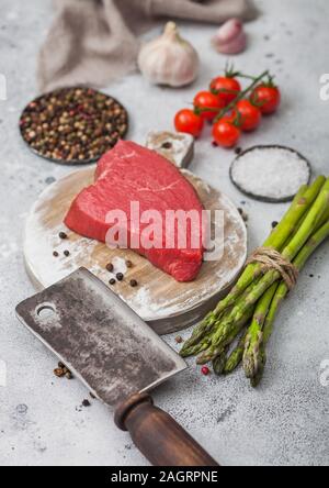 Tranche de boeuf cru sirlion steak sur planche à découper ronde avec des tomates, l'ail et d'asperges et de viande hache de table de cuisine lumière arrière-plan. Banque D'Images