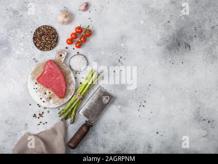 Tranche de boeuf cru sirlion steak sur planche à découper ronde avec des tomates, l'ail et d'asperges et de viande hache de table de cuisine lumière arrière-plan. Banque D'Images