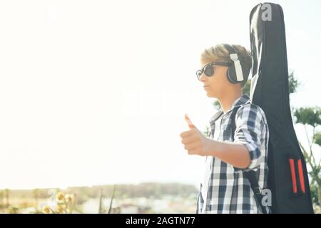 Essayez d'arrêter à la mode jeune garçon voiture pouce vers le haut. L'autostop adolescent avec guitare sur ses épaules. Voyageur Voyageur en attente d'amis. Par voyage autosto Banque D'Images