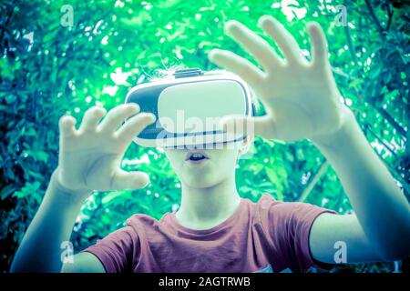 Jeune homme d'avoir du plaisir avec la technologie des lunettes de réalité virtuelle dans le parc en plein air. Garçon étonné gesticule portant casque vr Tech vie moderne Banque D'Images