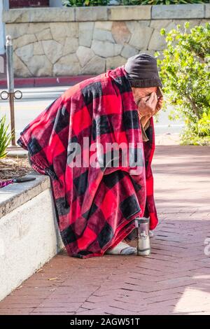 Une personne sans-abri est assis, penché au-dessus, sur un mur très bas par le trottoir de State Street à Santa Barbara, CA recouvert d'une couverture rouge et noir. Banque D'Images