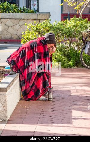 Une personne sans-abri est assis, penché au-dessus, sur un mur très bas par le trottoir de State Street à Santa Barbara, CA recouvert d'une couverture rouge et noir. Banque D'Images