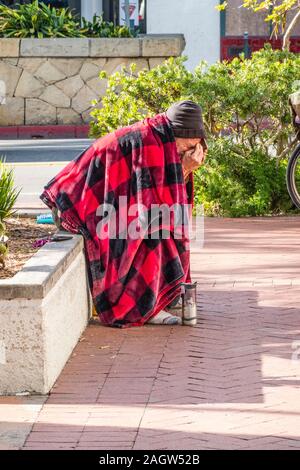 Une personne sans-abri est assis, penché au-dessus, sur un mur très bas par le trottoir de State Street à Santa Barbara, CA recouvert d'une couverture rouge et noir. Banque D'Images