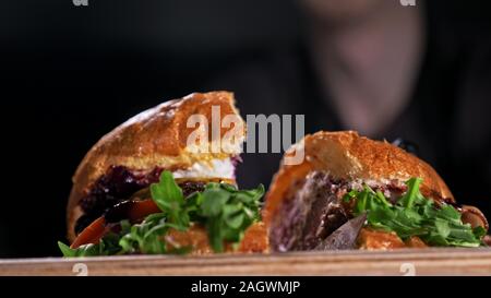 Pour couper la cuisson burger est sur fond noir. Composé : sauce, salade, tomate, oignon rouge, cornichon, fromage, bacon, pain et viande marbrée de boeuf. Banque D'Images