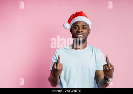 Drôle joyeux afro-américains non rasé jeune étudiant, tout sourire, wearing Santa hat, white t shirt, montrant le majeur des deux mains, isola Banque D'Images
