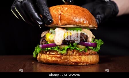 Burger d'artisanat est cuisson sur fond noir. Composé : sauce salsa, laitue, oignon rouge, cornichon, fromage, piment poivron vert, bun, marbré de bœuf viande. Banque D'Images