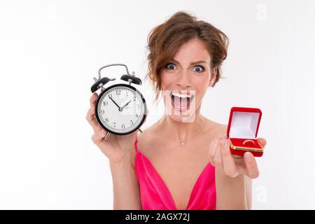 Fille charmante émotionnelle dans une robe rose avec une encolure est titulaire d'une boîte rouge avec un anneau de mariage et d'un réveil sur un fond blanc Banque D'Images