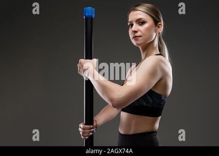 Strong girl in a sports top détient un bâton de gymnastique et se tient la moitié de face sur un fond sombre Banque D'Images