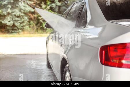 Côté de silver car lavés dans le libre service carwash, jet de pulvérisation d'eau sur le métal, le flou d'arrière-plan les arbres éclairés sun Banque D'Images