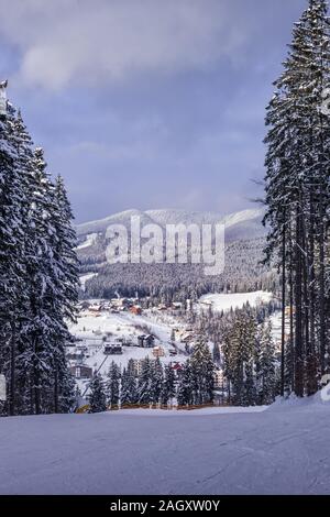 Vertical image avec vue sur le village de montagne de l'Ukraine comme vu à partir de la pente de ski entourée par de hauts arbres. Chaîne des Carpates à l'arrière-plan. Cloud Banque D'Images