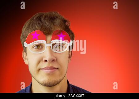 Portrait d'un homme à lunettes de Noël. Photo de smiling nerdy guy avec verres de Noël sur fond rouge. L'espace de copie pour le texte. Banque D'Images