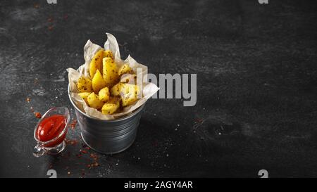 Quartiers de pommes de terre au four dans un seau en métal avec des herbes et de la sauce tomate sur fond noir - pommes de terre Légumes végétarien bio faits maison prêt à manger sn Banque D'Images