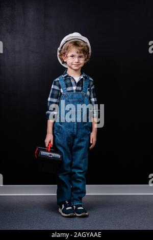 Adorable petit garçon en salopette en denim, casque et lunettes qui maintient la boîte à outils Banque D'Images