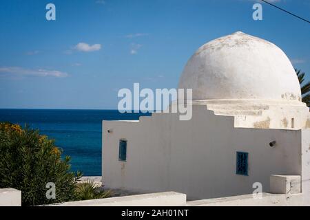 Un bâtiment peint en blanc avec des fenêtres bleues situé à côté d'un vieux cimetière avec des tombes blanches parmi les arbres au bord de la mer à Hergla, Tunisie Banque D'Images