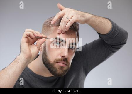 Élégant Studio portrait of young man standing against gray wall background façonner ses sourcils à l'aide de pincettes Banque D'Images