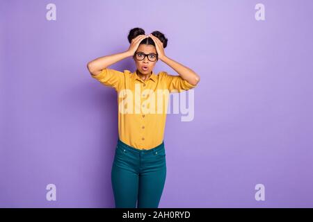 Photo de la peau sombre assez étonnante dame tenant les mains sur la tête pas terrible nouvelle, bouche ouverte, porter des specs chemise pantalon jaune pourpre isolés Banque D'Images