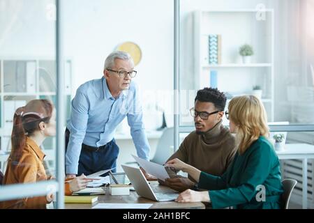 Mature businessman dans lunettes et permanent expliquant la stratégie d'entreprise pour les jeunes gens d'affaires à la table à l'office à la réunion Banque D'Images