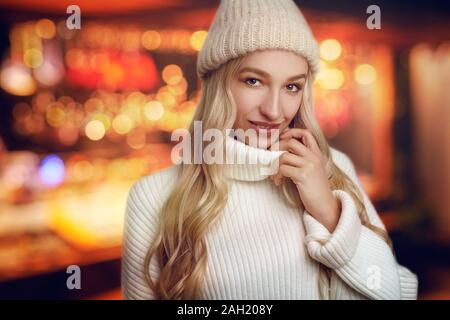 Très belle jeune femme blonde en blanc chandail tricoté et winter hat, looking at camera and smiling, debout contre les feux jaune brouillé avec b Banque D'Images