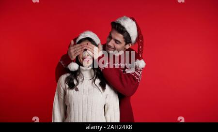 L'homme ferme les yeux petite amie avant sa surprise. Couple in Santa hat sur rouge Banque D'Images