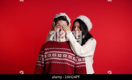 Ferme les yeux de femme petit ami avant de lui faire une surprise. Couple in Santa hat sur rouge Banque D'Images