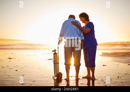 Infirmière réconfortant a senior male patient debout avec son réservoir d'oxygène sur une plage au coucher du soleil. Banque D'Images