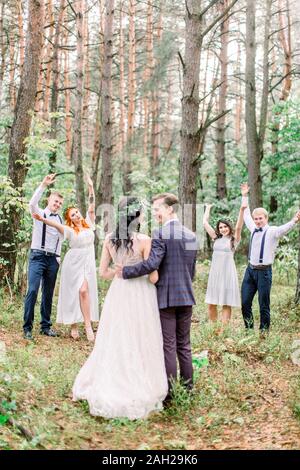 Couple de mariage rustique élégant avec des demoiselles et garçons d'honneur à forest sur jour de mariage. Vue arrière de l'époux et épouse, embrassant et en l'embrassant. Leur frie Banque D'Images