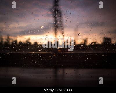 La silhouette floue de l'environnement industriel à travers le verre de voiture avec des gouttes de pluie contre la fenêtre. Coucher du soleil et nuages en arrière-plan Banque D'Images