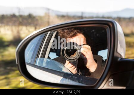 Photographe masculin reflété dans le miroir latéral de la voiture de prendre des photos avec appareil photo à travers la fenêtre ouverte tout en voyageant dans la campagne en été Banque D'Images