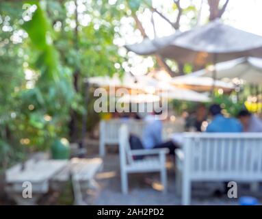 Plein air sur l'arrière-plan flou cafe journée ensoleillée. Flou des gens assis sur chaise en bois blanc sous parapluie dans le jardin public doté d'arbre vert. Banque D'Images