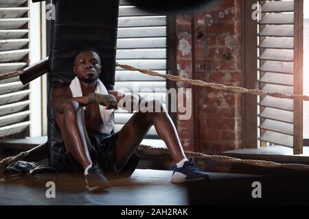 Portrait of African boxer musculaire looking at camera, assis sur le sol et se reposant après un entraînement sportif Banque D'Images