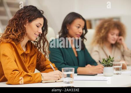 Niveau horizontal tourné de trois jeunes femmes élégantes prendre des notes au cours de réunion d'affaires dans un bureau moderne