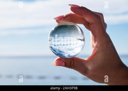 La main de femme tenant une boule de cristal, à la recherche jusqu'à l'océan et le ciel. La photographie créative, crystal ball réfraction Banque D'Images