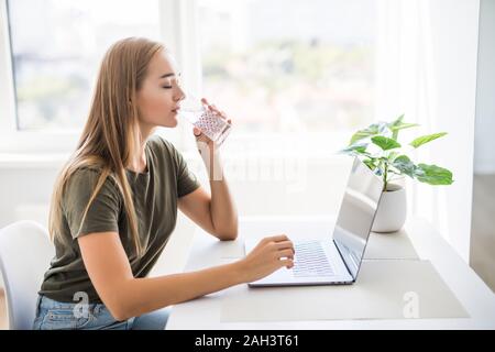 Femme buvant de l'eau tout en tapant de verre à son ordinateur portable. Banque D'Images