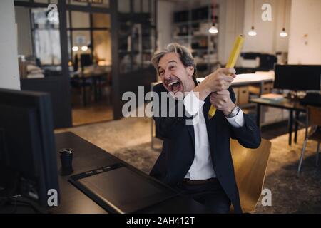 Angry senior businessman avec crayon géant at desk in office Banque D'Images