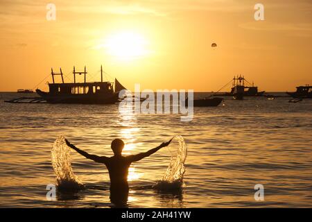 Silhouette of man at beach les projections d'eau avec les bras grand ouverts et voiliers au coucher du soleil à White Beach Boracay, Philippines. Vacances d concept Banque D'Images