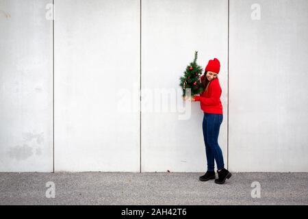 Woman wearing red pull et chapeau, wolly holding arbre de Noël artificiel devant un mur Banque D'Images